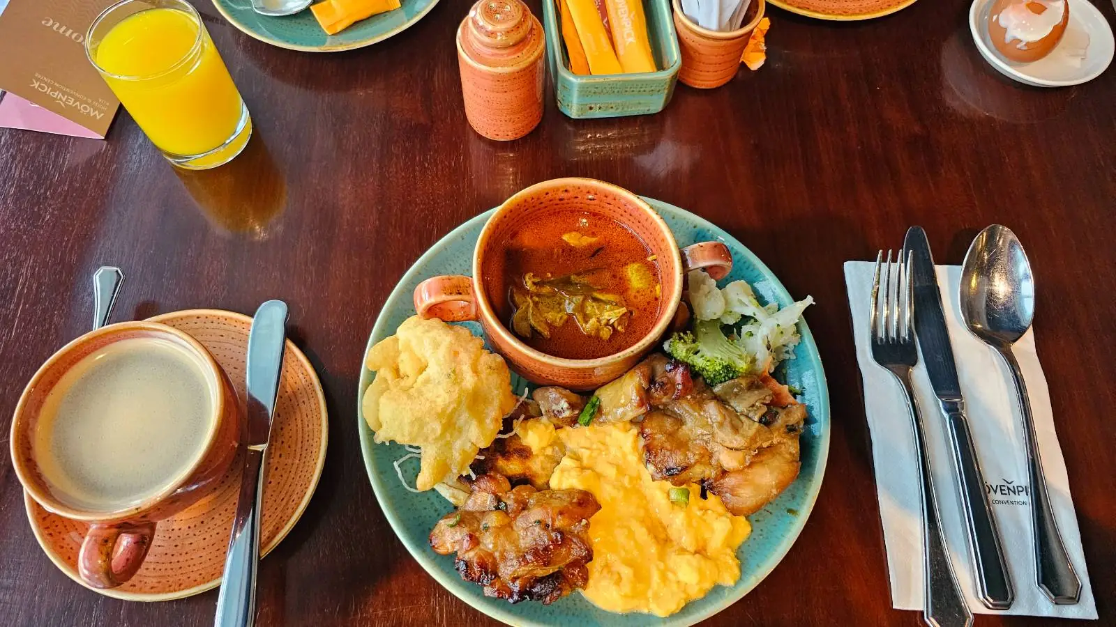 A hearty breakfast spread on a wooden table features a plate with scrambled eggs, fried chicken, mashed potatoes, and a small salad. A bowl of soup sits on the plate, while a cup of coffee, a glass of orange juice, and cutlery are neatly arranged nearby.