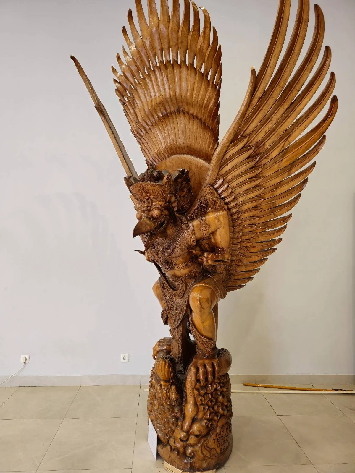 Colorful statue of Garuda, a mythical bird figure, with large red and blue wings, intricate carvings, and vibrant details. The statue is placed on a pedestal in a gallery setting with a plant visible to the side.