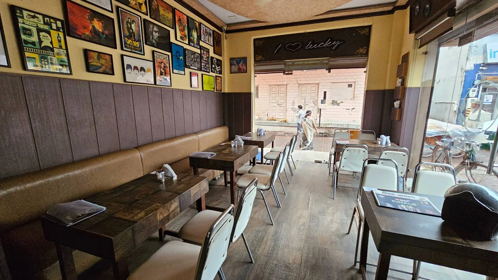 A cozy cafe interior with wooden tables and white chairs lined against one wall. Framed colorful artwork decorates the walls. The cafe is empty, and a large window at the end shows a blurred outdoor scene.