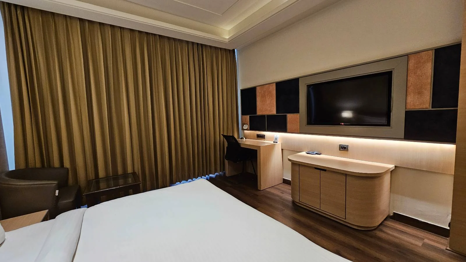 Modern hotel room with a large bed, desk and chair, flat-screen TV, armchair, wooden furnishings, and floor-to-ceiling brown curtains covering the window.