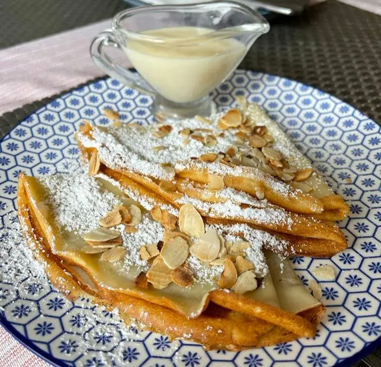 Crepes topped with almonds accompanied by a clear glass of condensed milk.