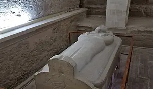 A stone sarcophagus is displayed within a fenced area in a dimly lit, ancient, and worn stone room.