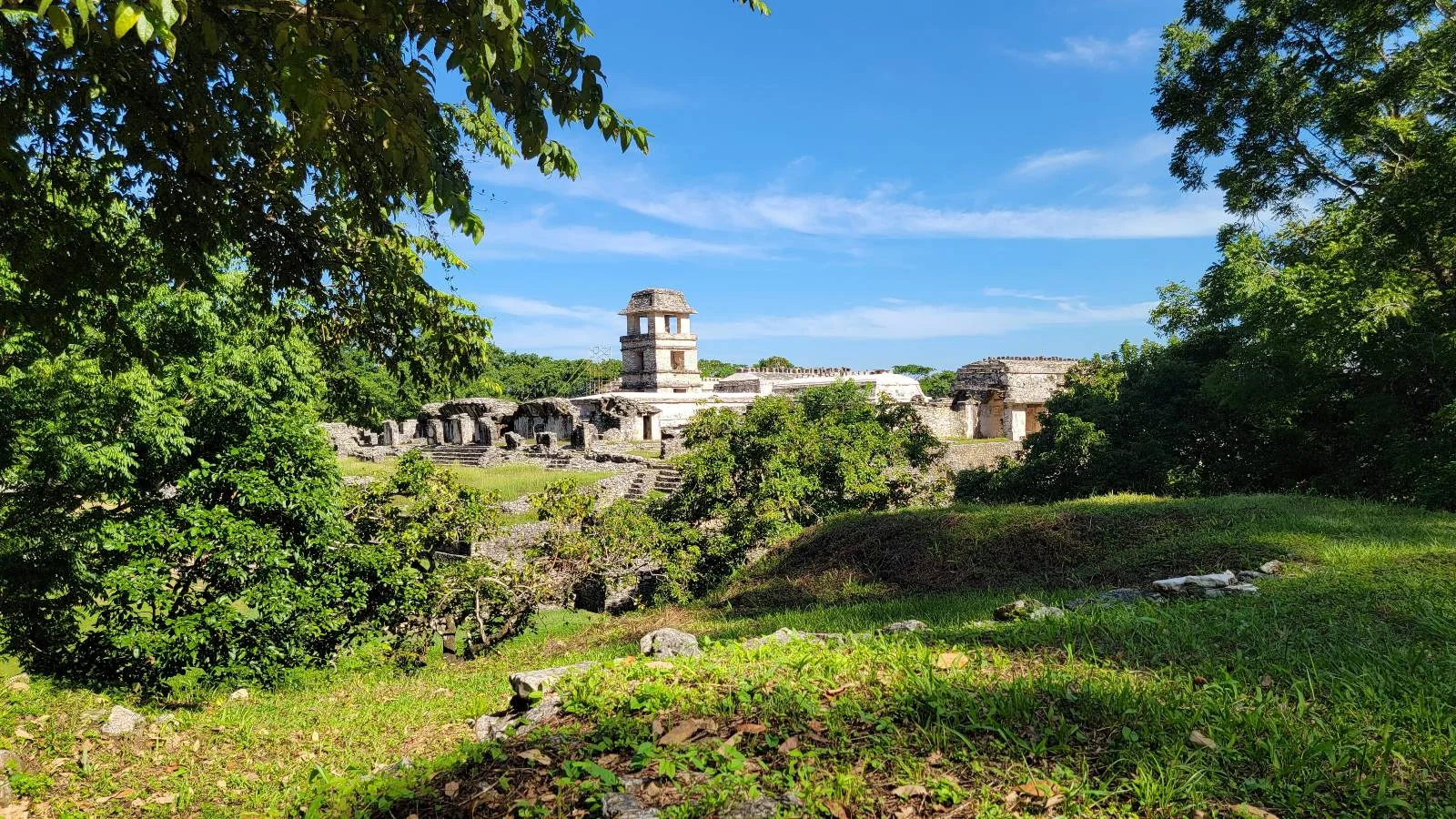 View of a historic ancient temple complex surrounded by lush greenery under a clear blue sky. The central structure is prominent against the natural landscape, with trees and grass framing the scene.