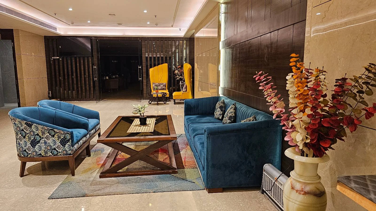 A modern indoor seating area with a blue couch, patterned chair, glass coffee table, colorful rug, a decorative statue, a floor lamp, and a red patterned pouf on a tiled floor near tall windows and wooden blinds.