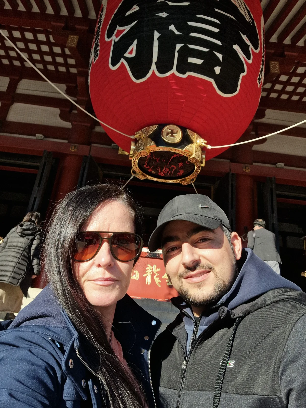 Two people smile beneath a large red lantern with black kanji, situated at a temple entrance. Bright sunlight, traditional setting.