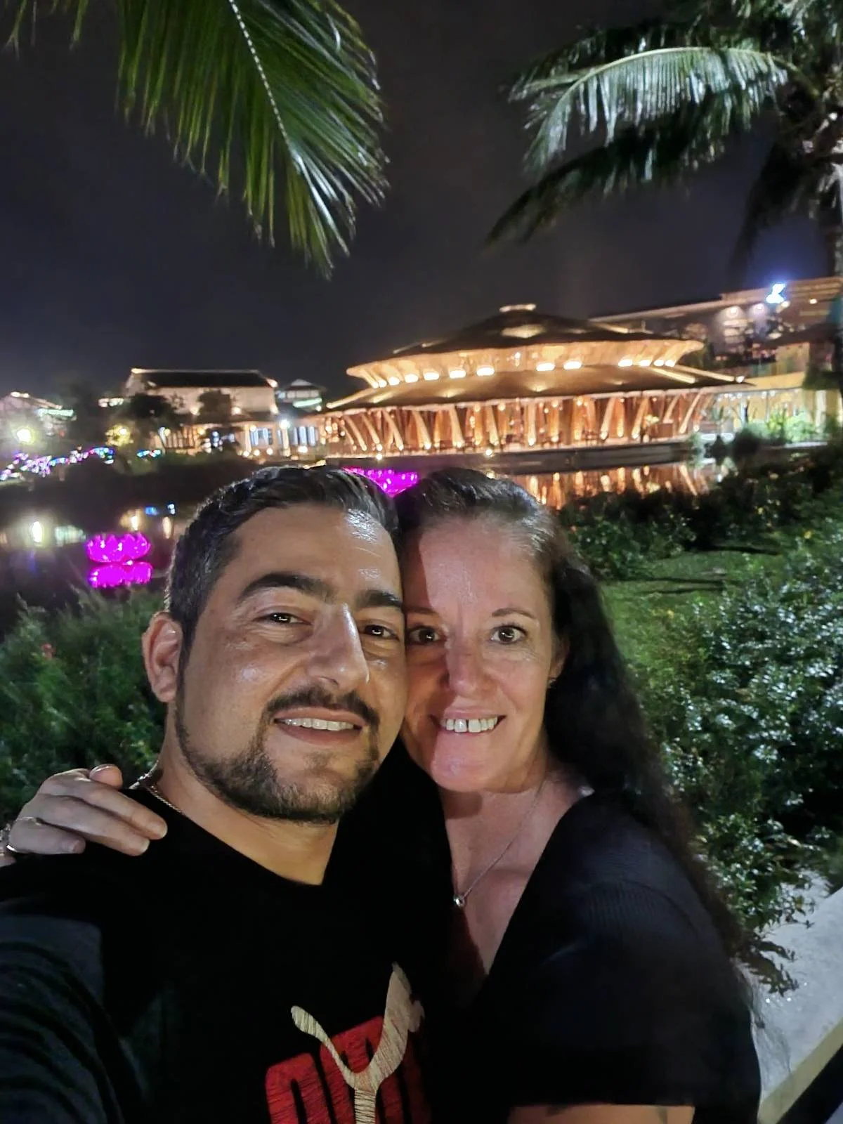 A couple posing for a selfie at night with illuminated buildings and palm trees in the background.