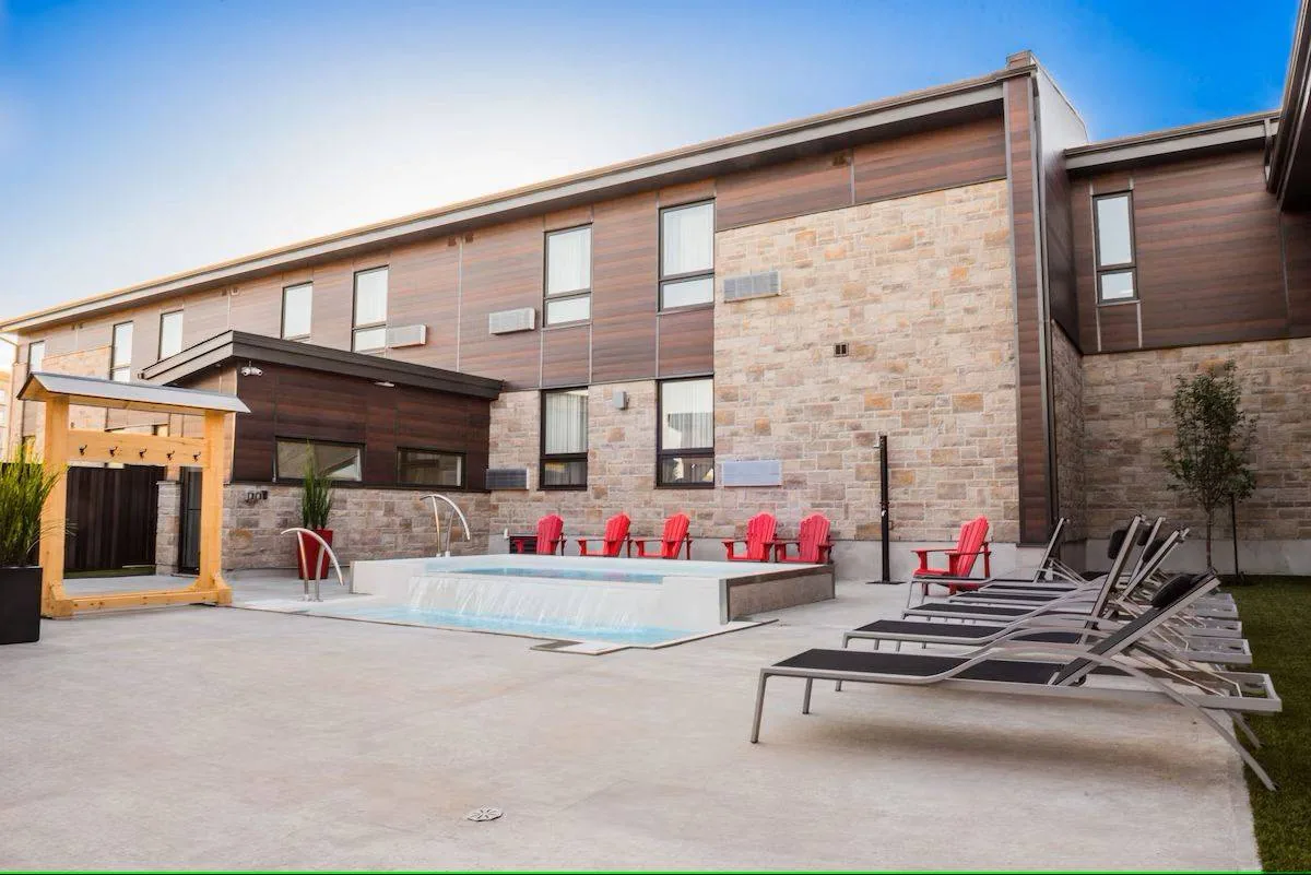 A modern building with a mix of stone and wood exterior features a small rectangular pool surrounded by red Adirondack chairs. There are several loungers on the patio, and the sky is clear and blue.