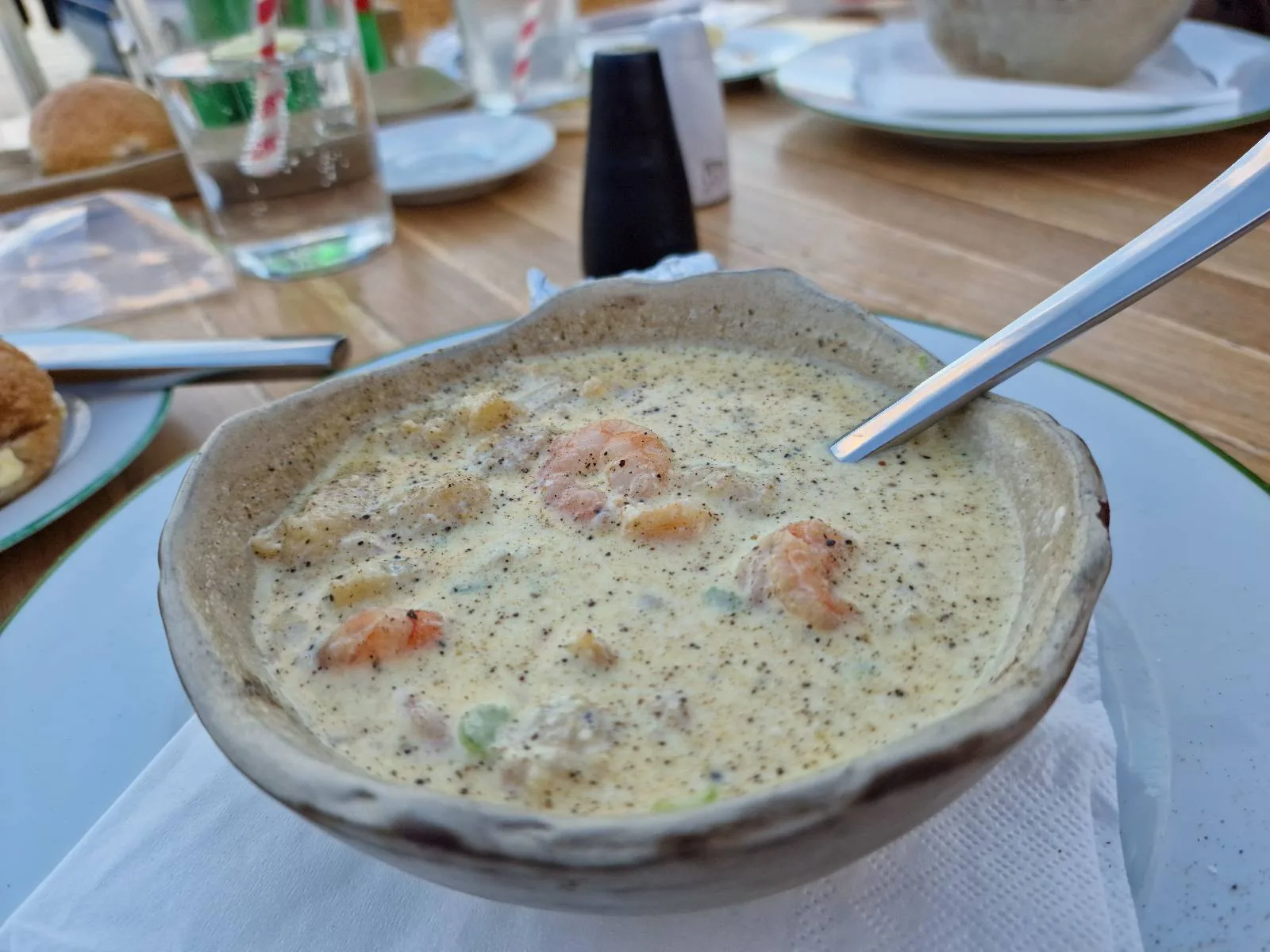 A bowl of creamy seafood chowder with shrimp and other ingredients is on a table with cutlery and glasses in the background.