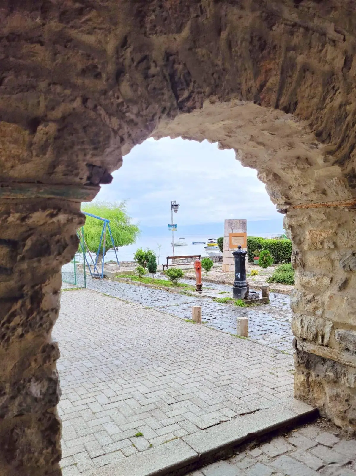 View through a stone archway showing a cobblestone path leading to a waterfront area. A lamppost and a few people are visible near the water, with trees and a cloudy sky in the background.