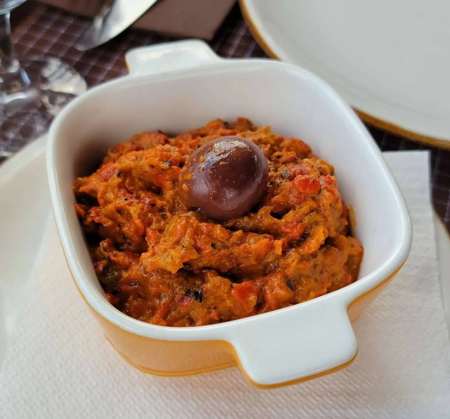 A square white dish filled with a chunky red and brown spread, possibly a roasted eggplant or pepper dip, garnished with a single dark olive. The dish rests on a white napkin beside a set table.