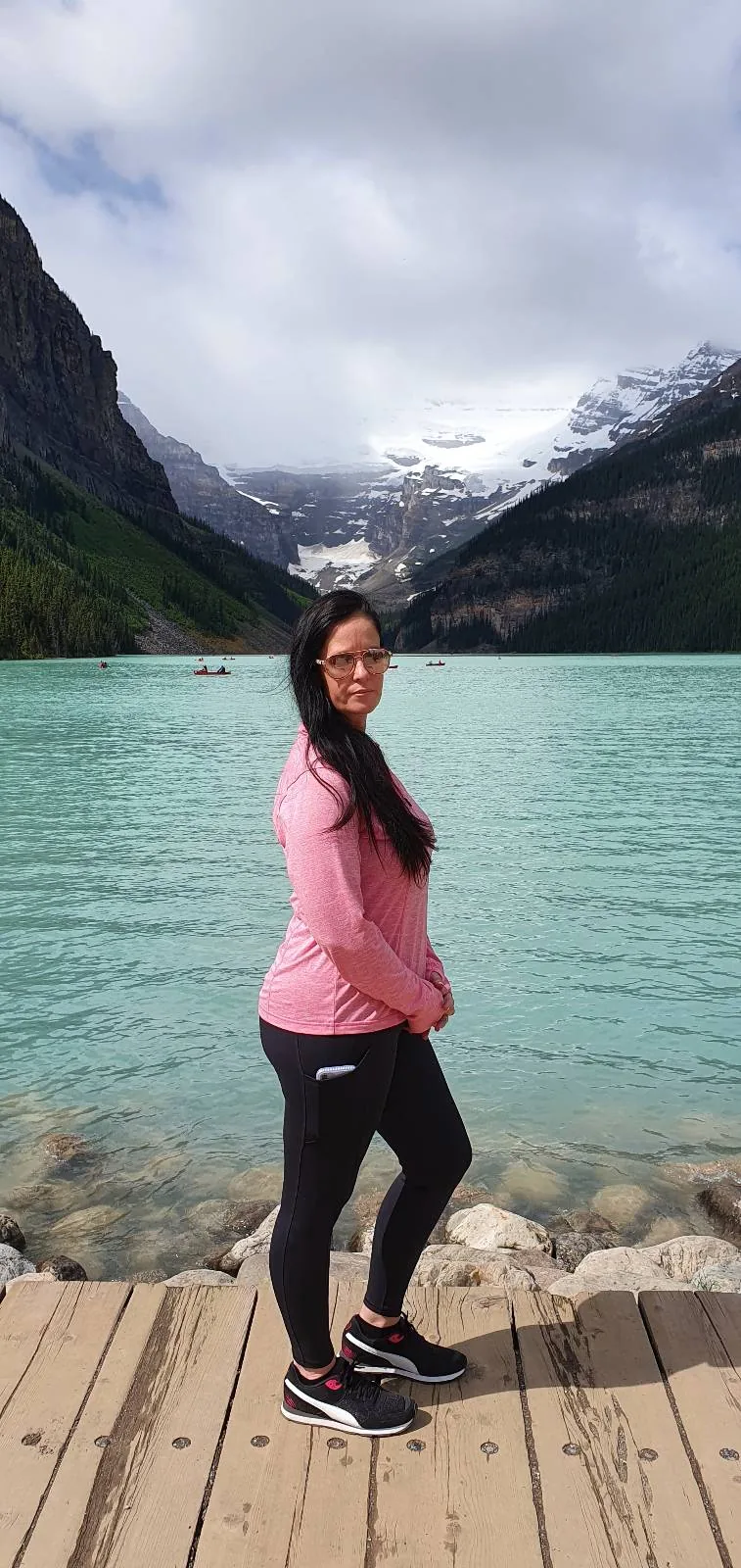 A woman in a pink jacket and black pants stands on a wooden dock by a turquoise lake, with mountains and cloudy sky in the background.