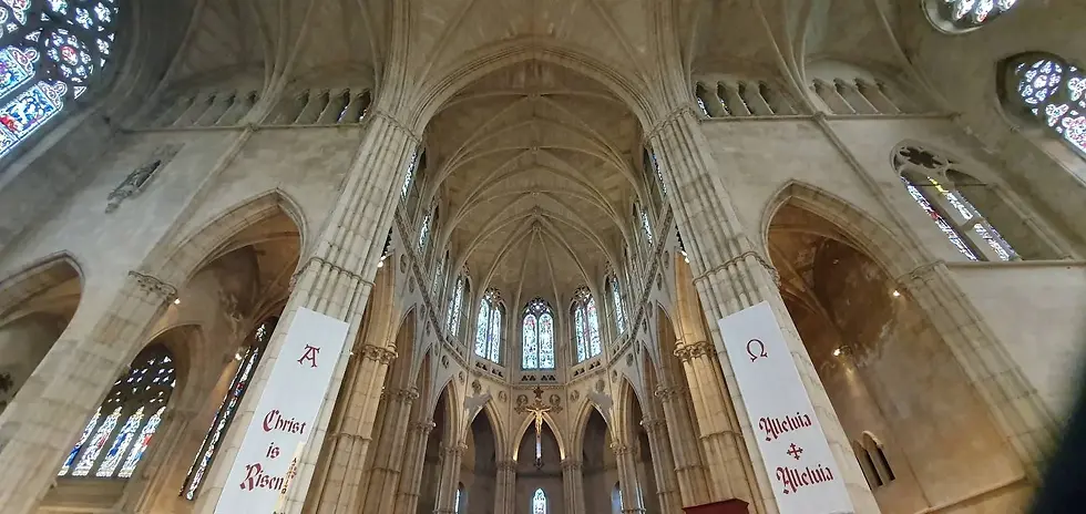 Gothic church interior with vaulted ceiling. Stained glass windows and banners reading "Christ is Risen" and "Alleluia" in red. Peaceful ambiance.