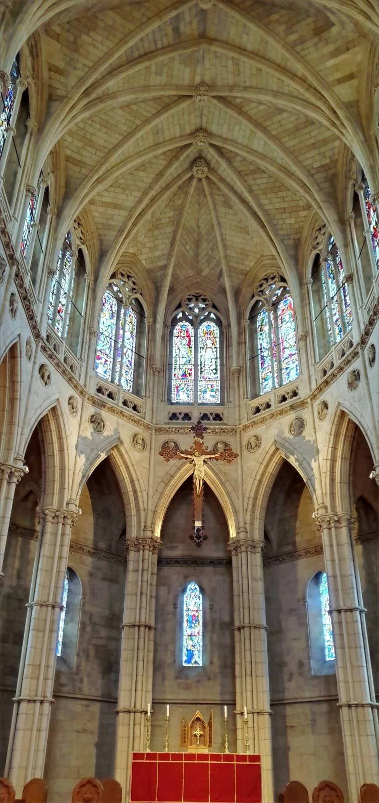 A view of a cathedral's ornate interior, showcasing a vaulted ceiling with intricate designs, arched windows with stained glass, and stone columns. A red altar sits at the center against a backdrop of tall, narrow windows.