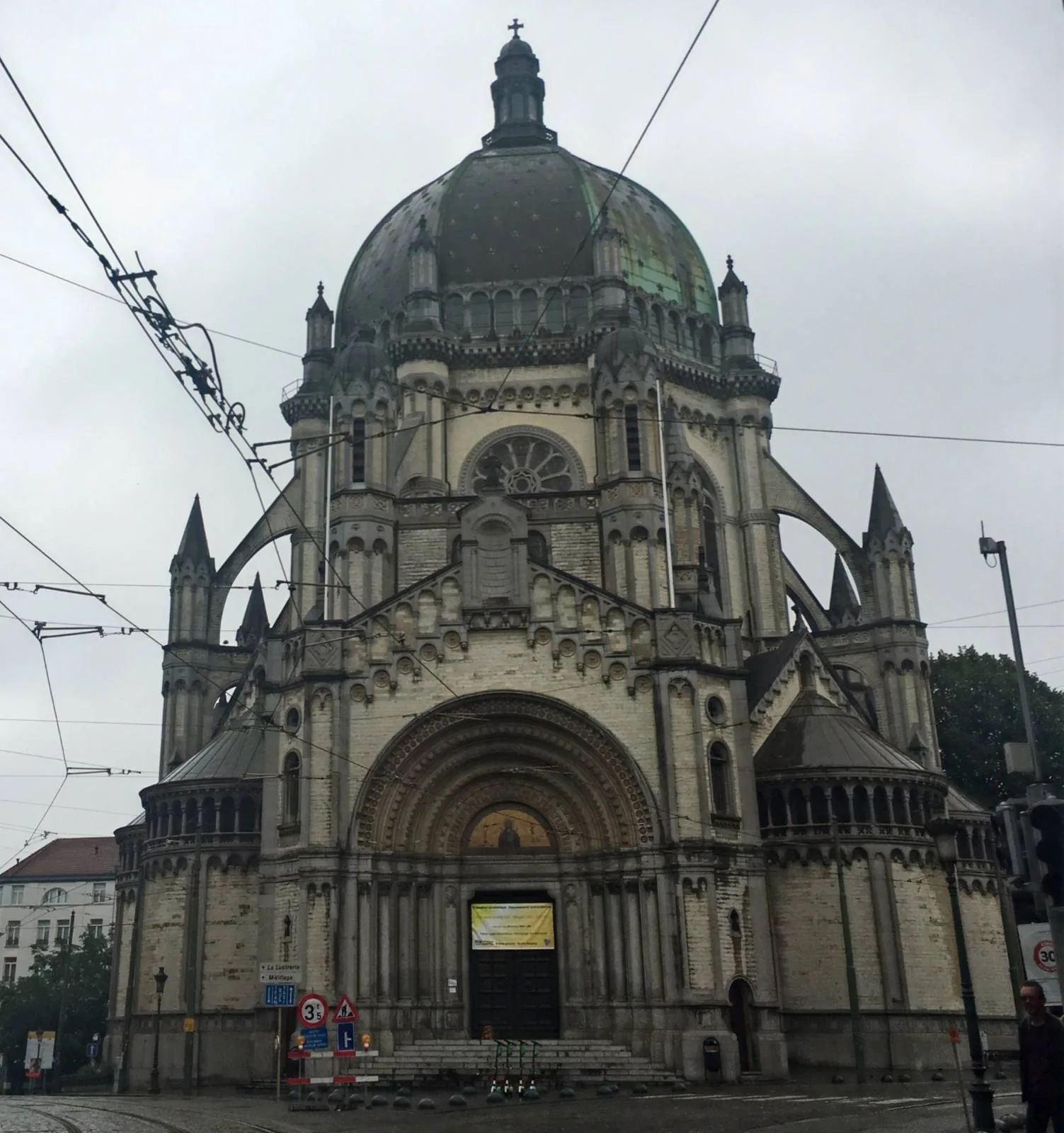 A large, ornate church with a prominent central dome and multiple spires, set against a cloudy sky. The architecture features detailed arches and columns. Tram lines and a traffic sign are visible in the foreground.
