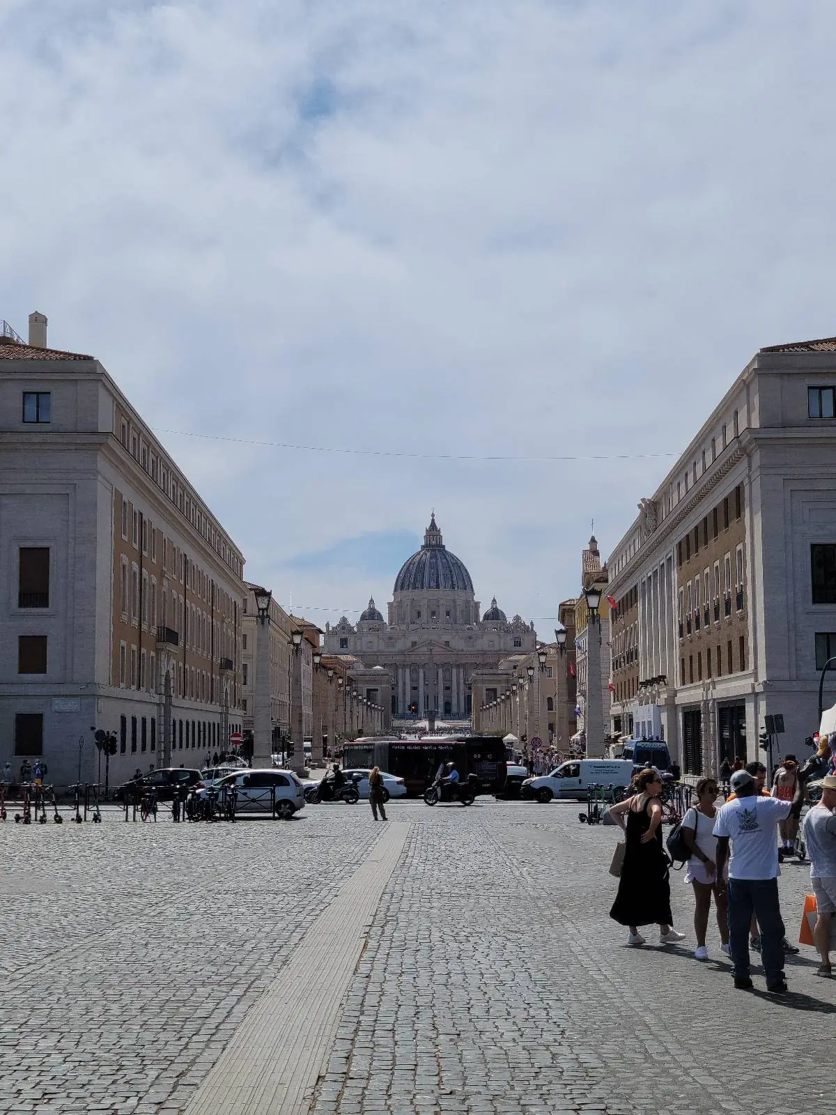 A wide cobblestone street lined with buildings leads to St. Peter’s Basilica in Vatican City, with people walking and cars parked along the sides under a partly cloudy sky.