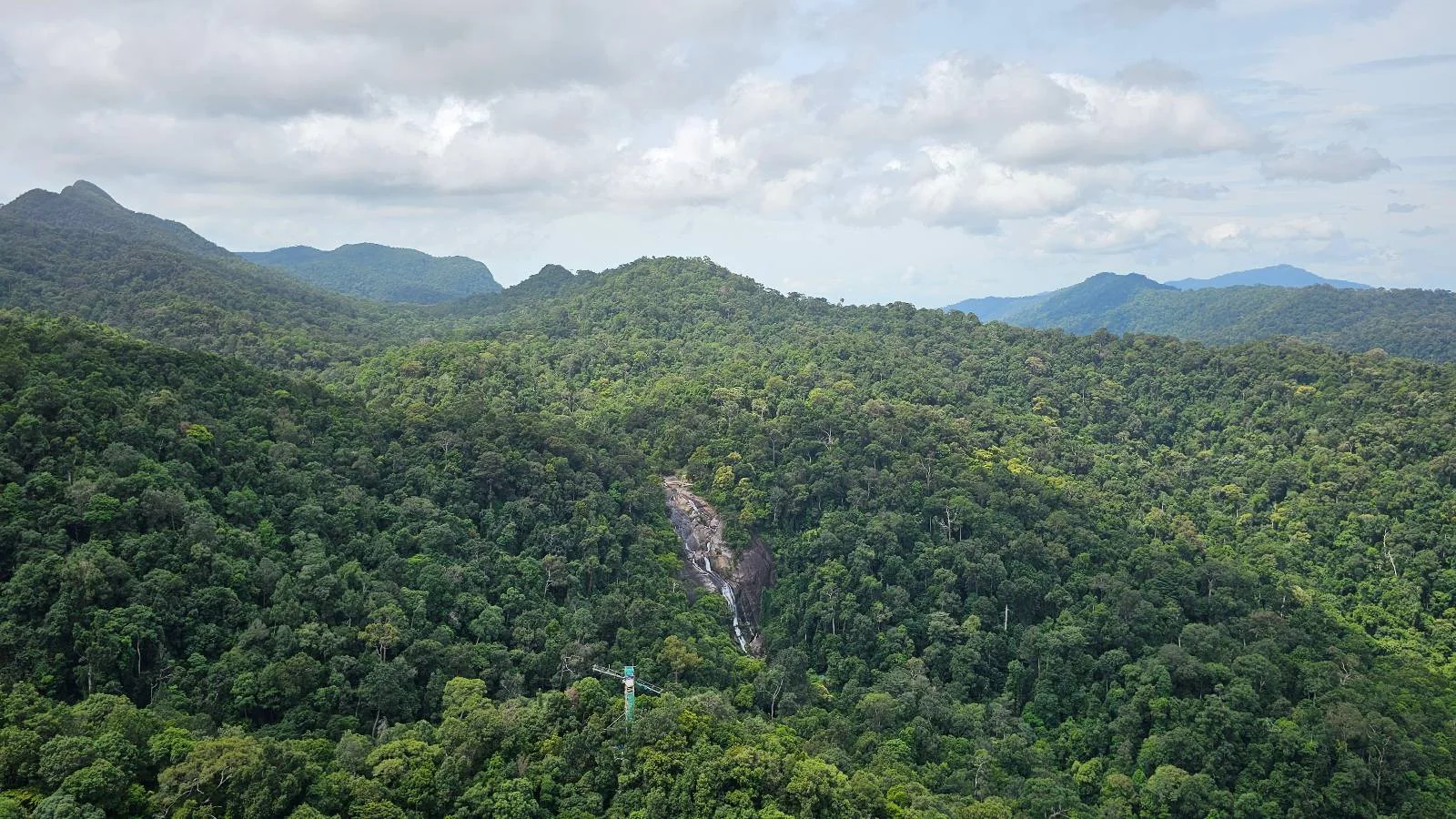 Aerial view of a lush, green forested landscape with rolling hills. A narrow river runs through the middle, surrounded by dense trees. The sky is overcast with patches of white clouds.