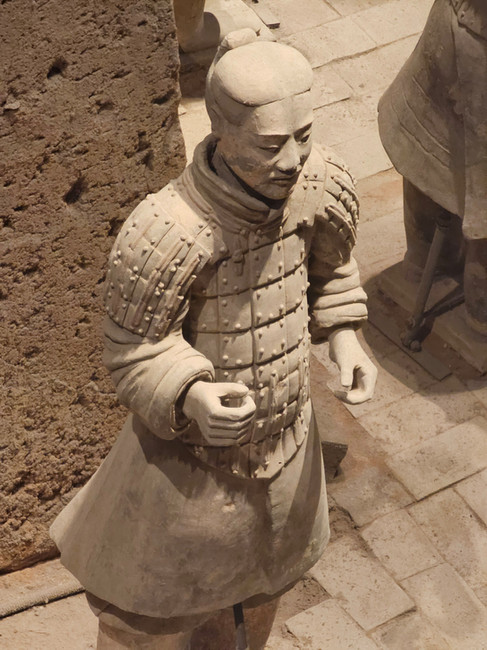 A clay statue of a Terracotta Warrior stands upright, wearing detailed armor with a topknot hairstyle, viewed from above. The figure holds one hand in a fist and the other in a partial grasp.
