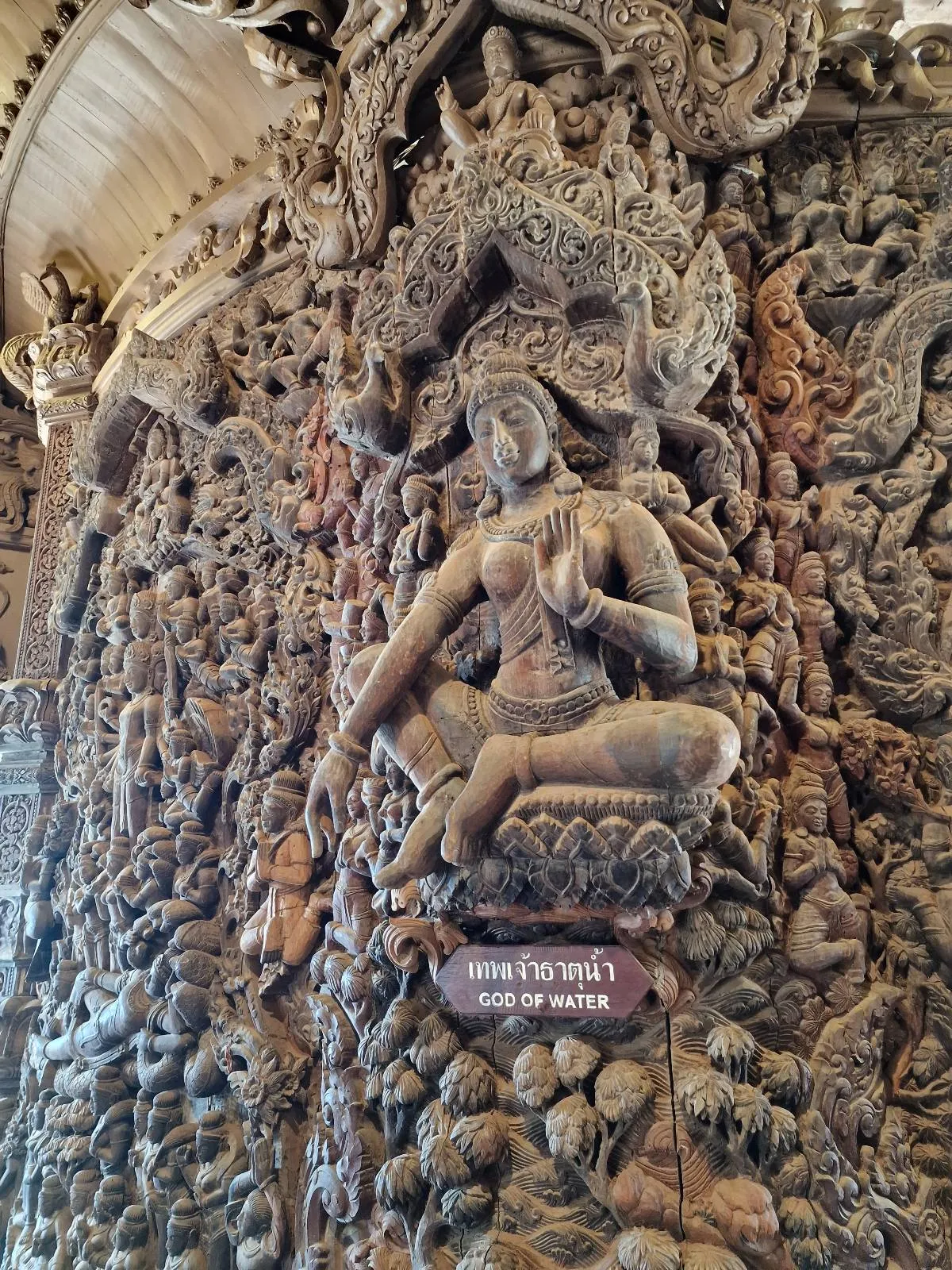 Intricate wooden carving depicting a seated figure surrounded by elaborate patterns and mythical creatures. The artwork is detailed and ornate, showcasing a rich display of craftsmanship. A small sign reads "God of Water.