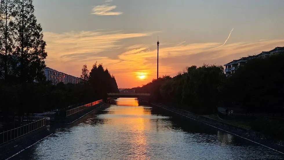 Sunset over a tranquil river with orange reflections. Trees and buildings line the riverbanks. Soft clouds linger in the sky.