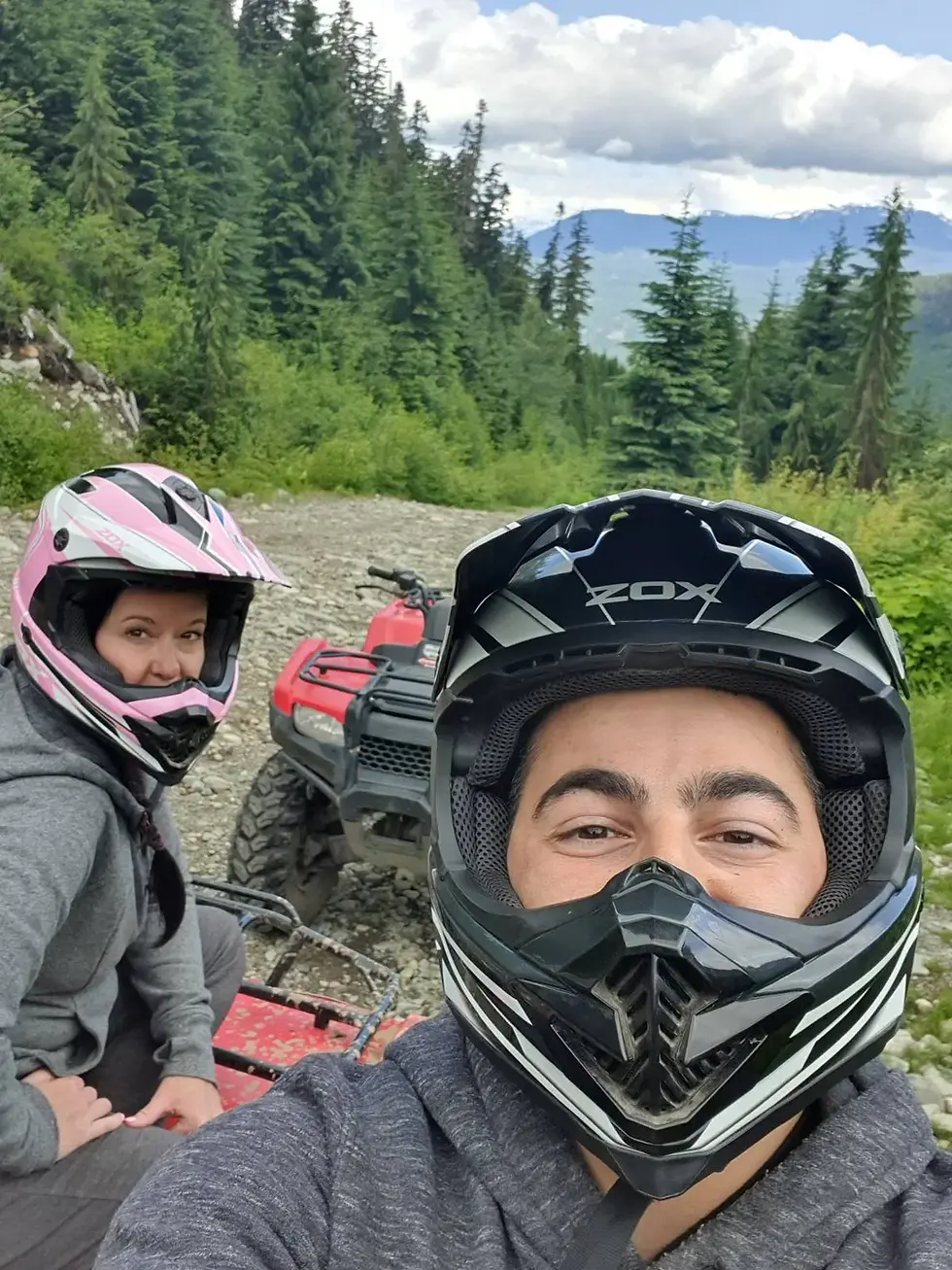 Two people wearing helmets are riding ATVs on a forested trail. The background features trees and mountains under a partly cloudy sky.