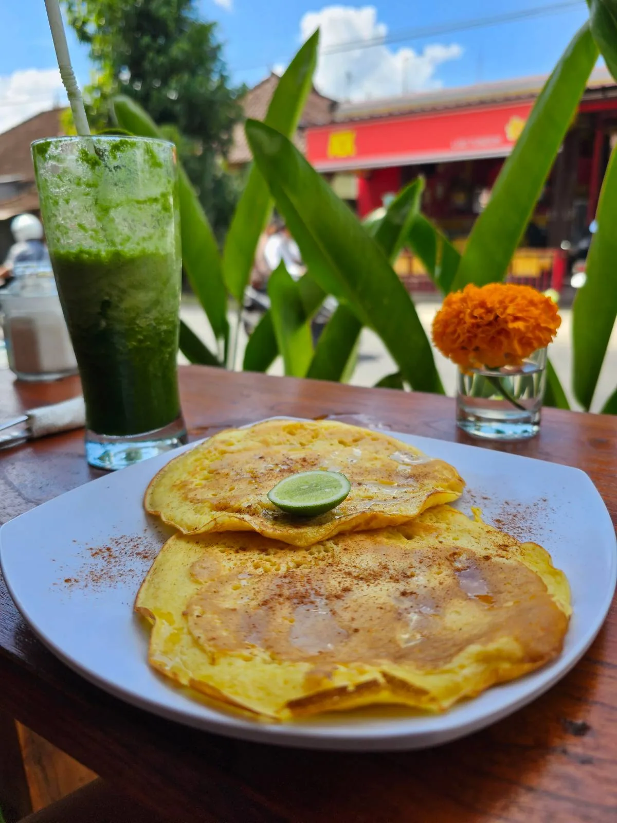 A close-up of two pancakes topped with a lime slice on a white plate. A tall glass of green juice, a small orange flower in a vase, and outdoor greenery are in the background on a sunny day.