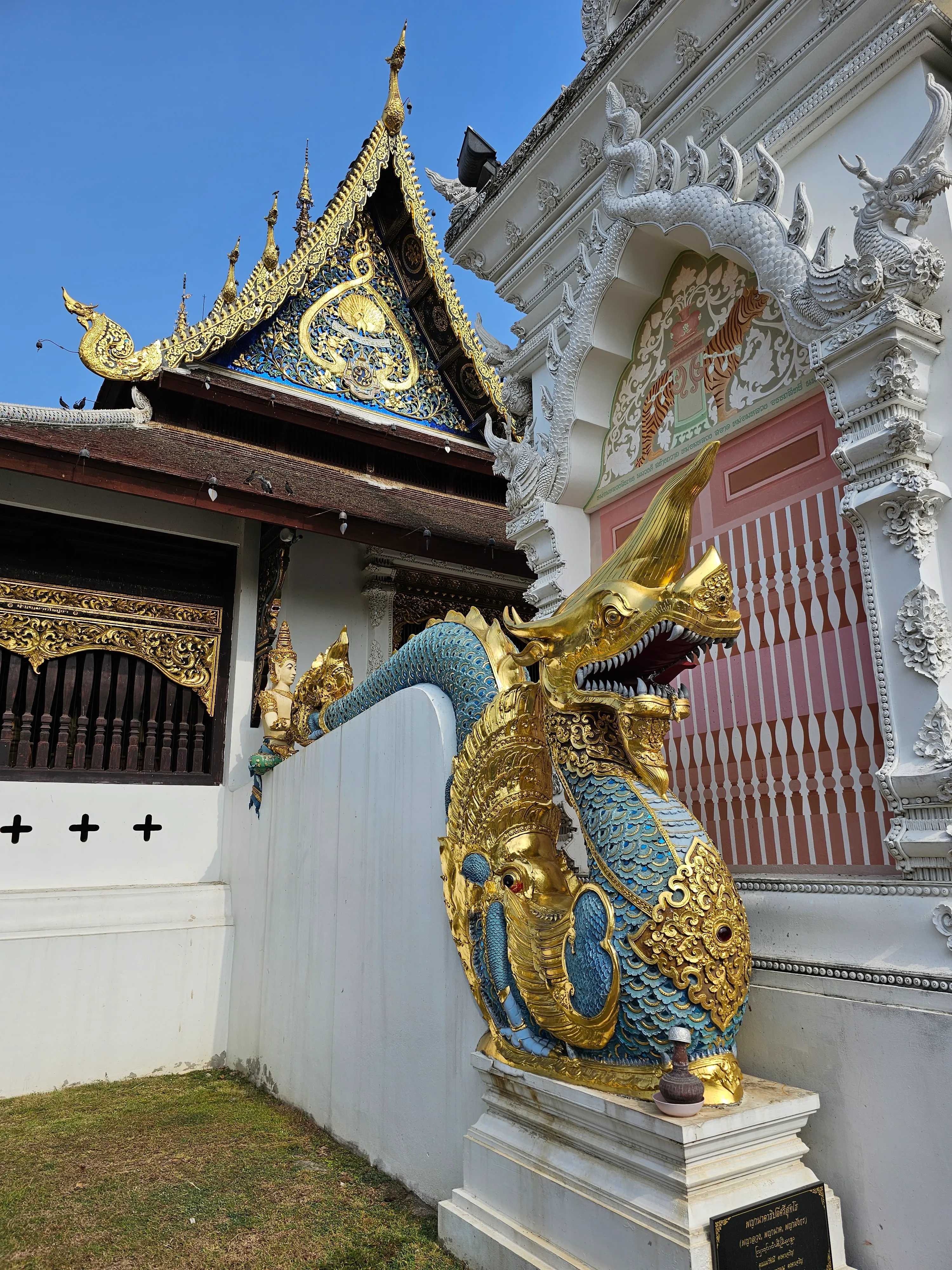 A traditional Thai temple features an ornate gold and blue dragon sculpture along a white wall, with intricate roof and architectural details visible in the background.
