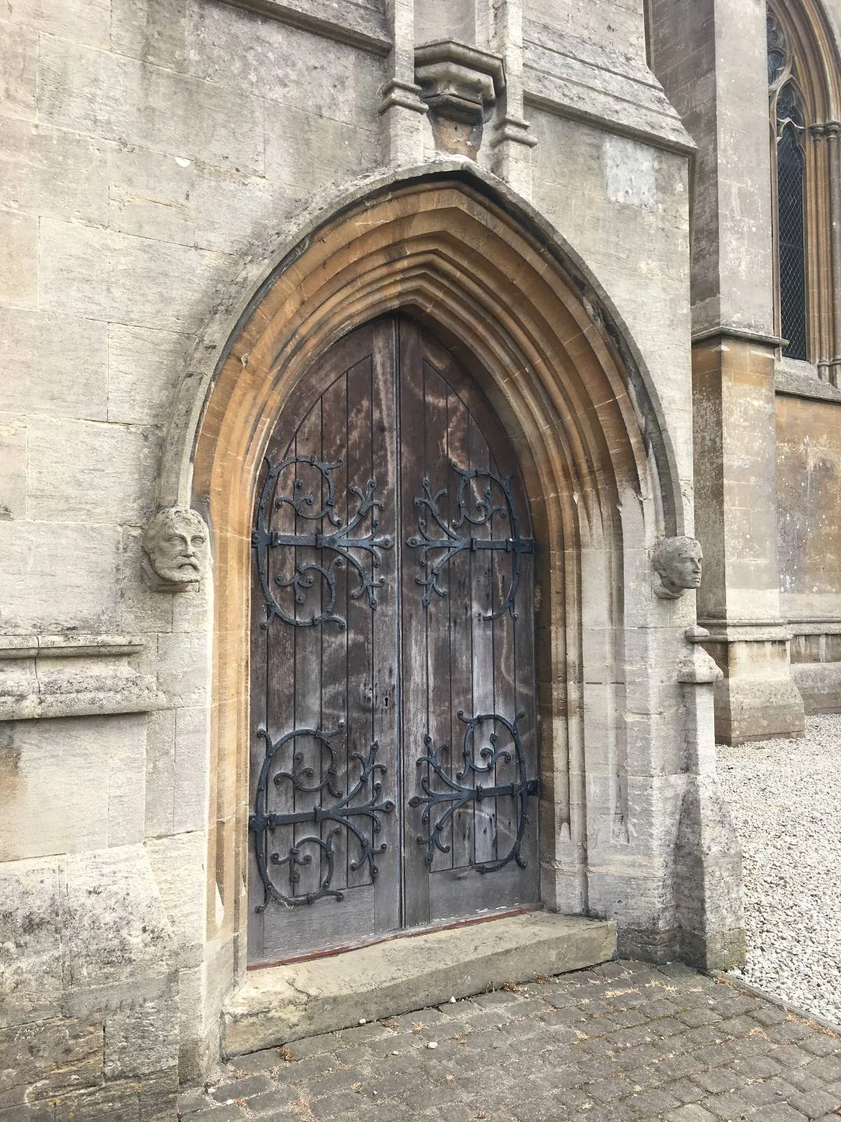 An ornate wooden door with intricate ironwork set within a pointed stone archway. The surrounding stone wall has detailed carvings, hinting at a historical or religious building. The ground is paved with cobblestones.