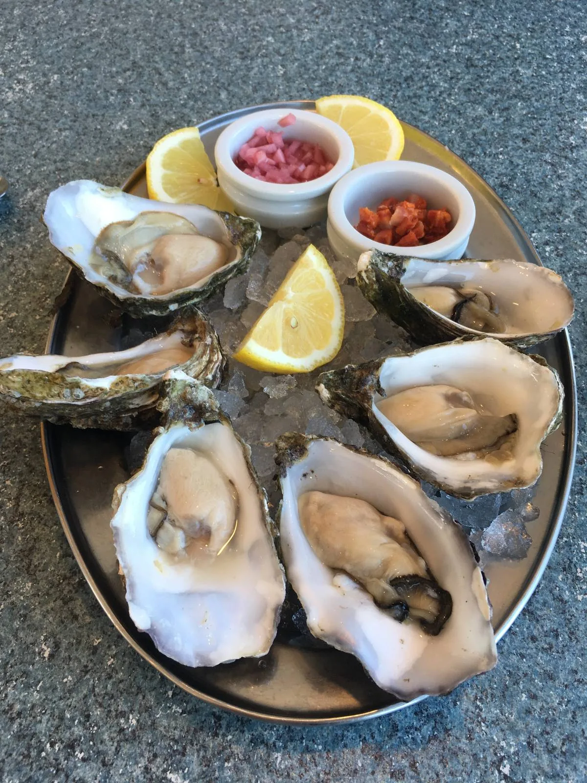 A platter of six oysters on the half shell is presented on a bed of ice. Lemon wedges and small containers of dipping sauces accompany the oysters, providing a fresh and appetizing seafood display.
