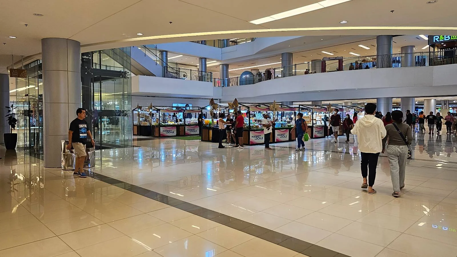 A spacious shopping mall interior with shiny floors and multiple levels. People are walking around, and a central kiosk with various stalls is visible. The architecture features modern design with glass and metal elements.