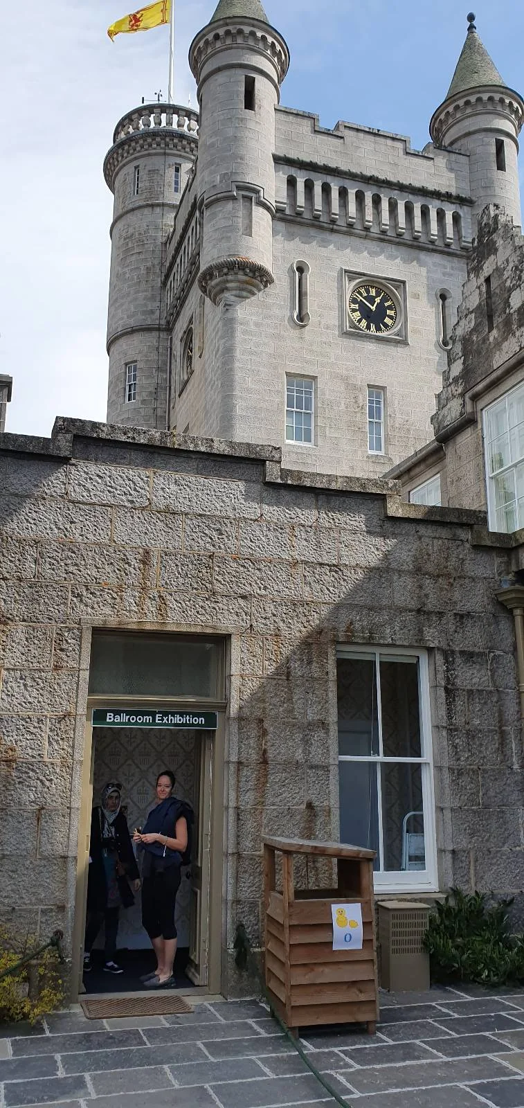 A person stands in the doorway of a stone building with turrets and a clock. There's a small wooden structure with a sign nearby. A flag flies atop one of the turrets under a partly cloudy sky.