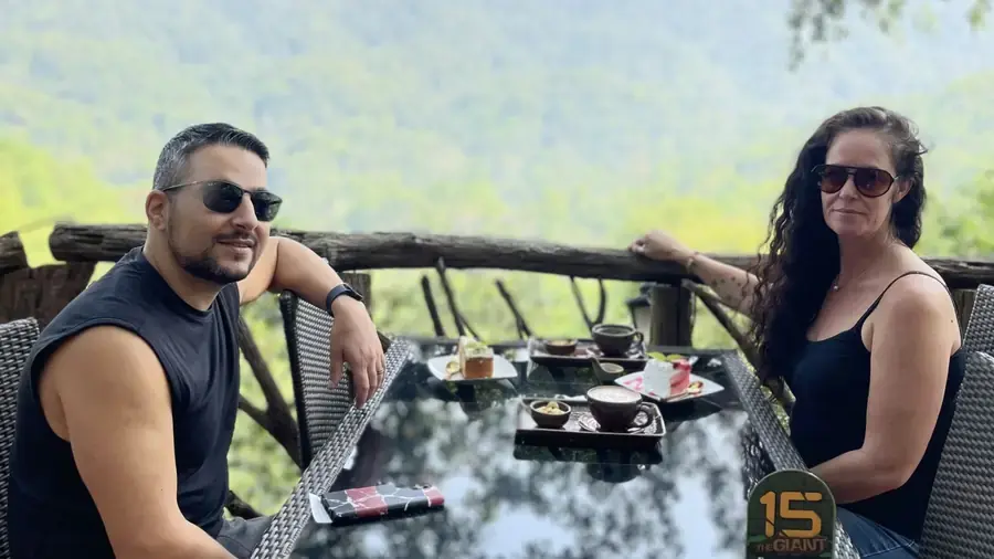 A man and woman sit at an outdoor table on a balcony at The Giant Chiang Mai, a unique treehouse café overlooking lush jungle views. Smiling in sunglasses, they enjoy a meal together with tall trees and mountains in the background.