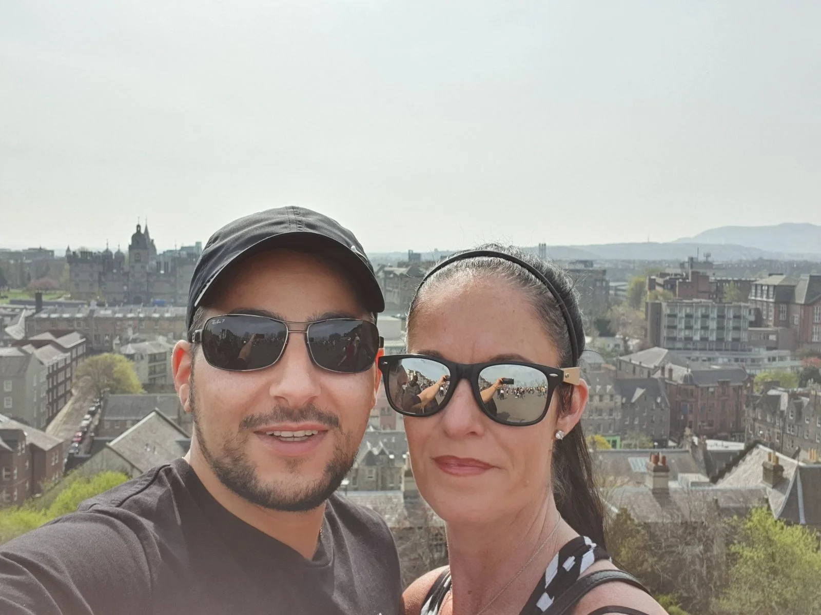 Jenn and Leon wearing sunglasses taking a selfie overlooking Edinburgh cityscape from a hillside viewpoint.