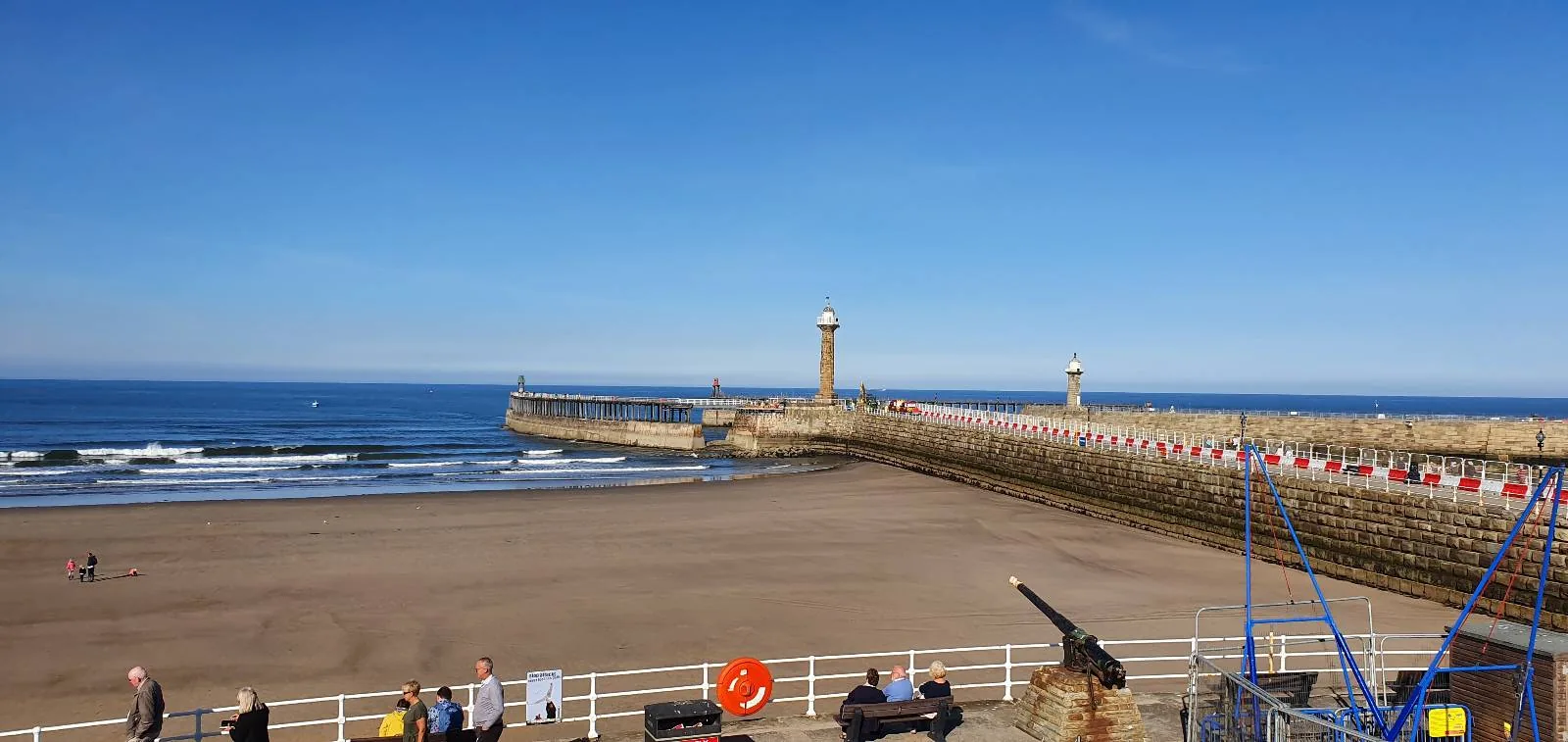 A sunny day at a sandy beach with a stone pier extending into the sea. Two lighthouses are visible on the pier. A few people are walking along the promenade, and a cannon is displayed on the pathway. The sky is clear and blue.