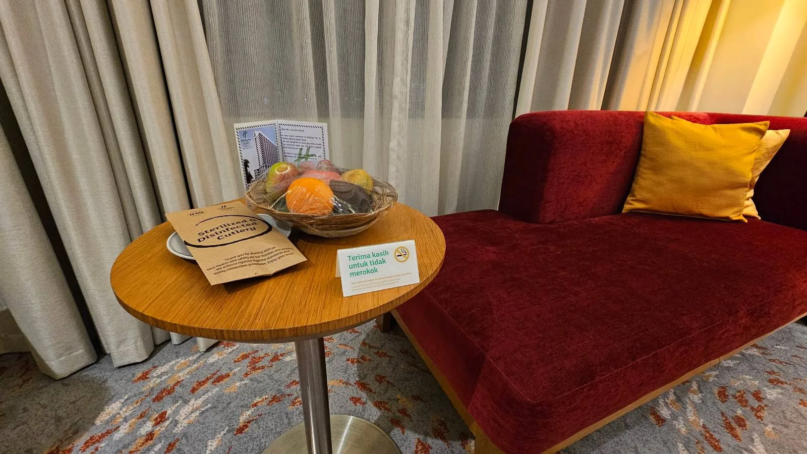 A small round table with a bowl of fruit, paper packets, and a card stands beside a red velvet chaise lounge with a yellow pillow; curtains hang in the background.