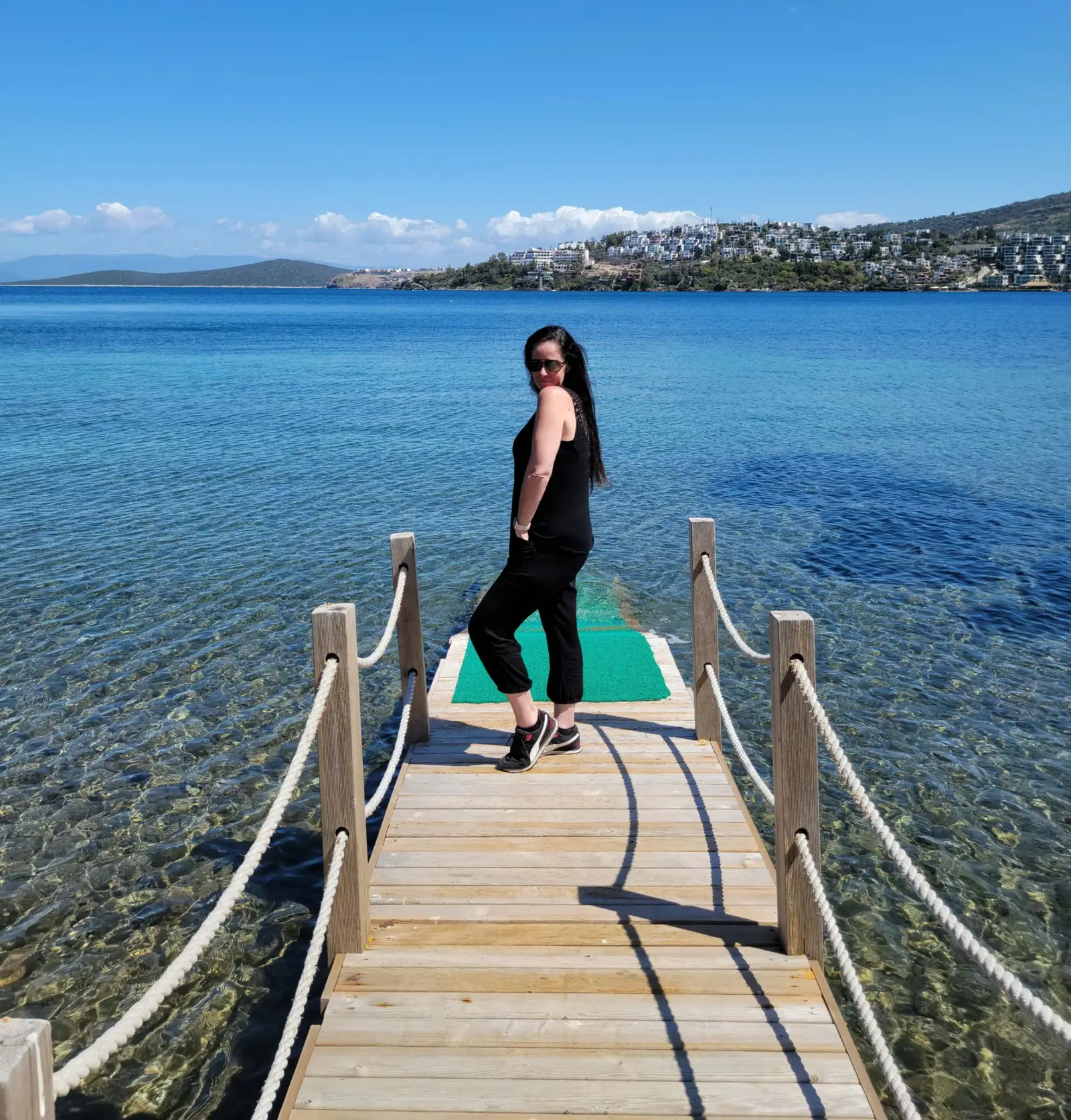 A woman in black clothing stands on a wooden pier over clear blue water, with a scenic coastal town and hills visible in the background under a bright, sunny sky.
