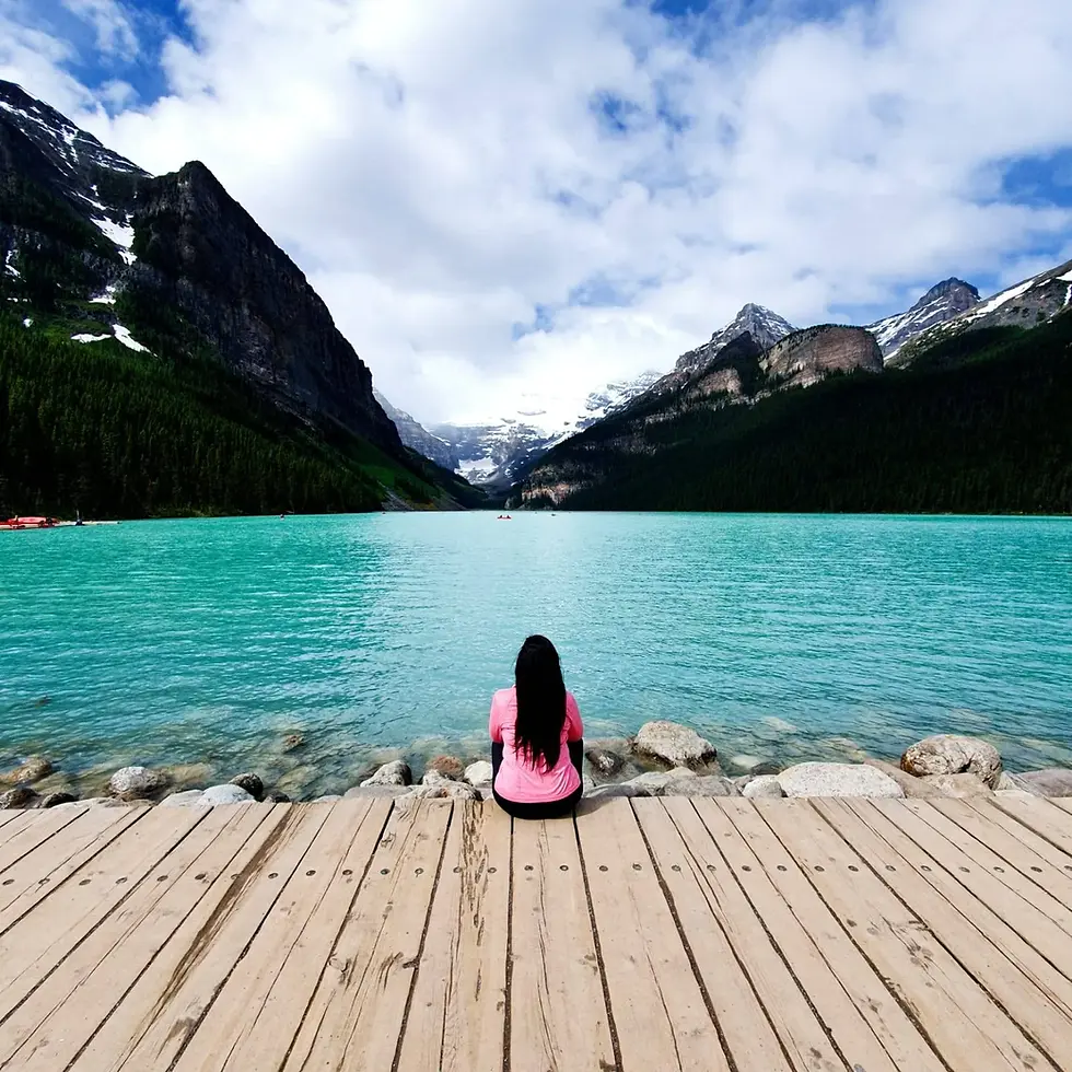 Person in a pink top sitting on a wooden dock, facing a turquoise lake surrounded by mountains under a cloudy sky.