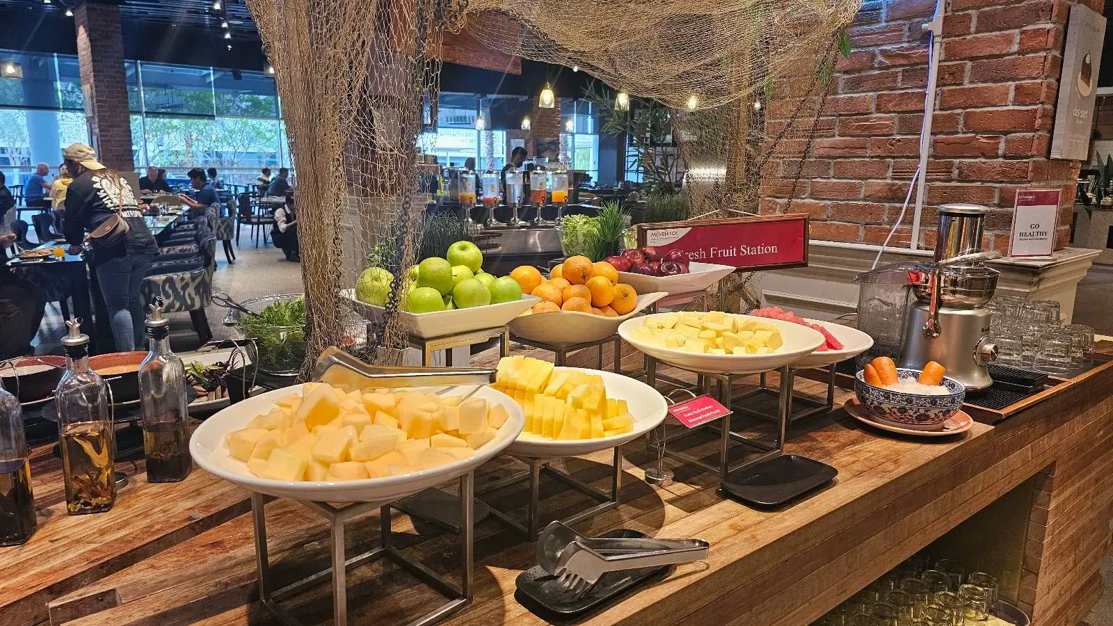 A vibrant buffet setup features bowls of fresh, sliced fruit including pineapples and oranges, with whole green apples and mandarins displayed in the background. The setting has a rustic feel with wooden surfaces and a brick wall, creating a welcoming atmosphere.