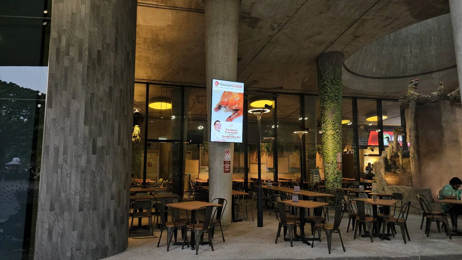 Outdoor seating area of a modern cafe with wooden tables and chairs. Large glass windows reveal the interior, and a digital sign is displayed on a column. The lighting is warm and inviting, and trees are visible in the background.