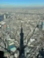 Aerial view of a cityscape with a large tower's shadow cast over buildings. Clear blue sky and rivers visible in the background.