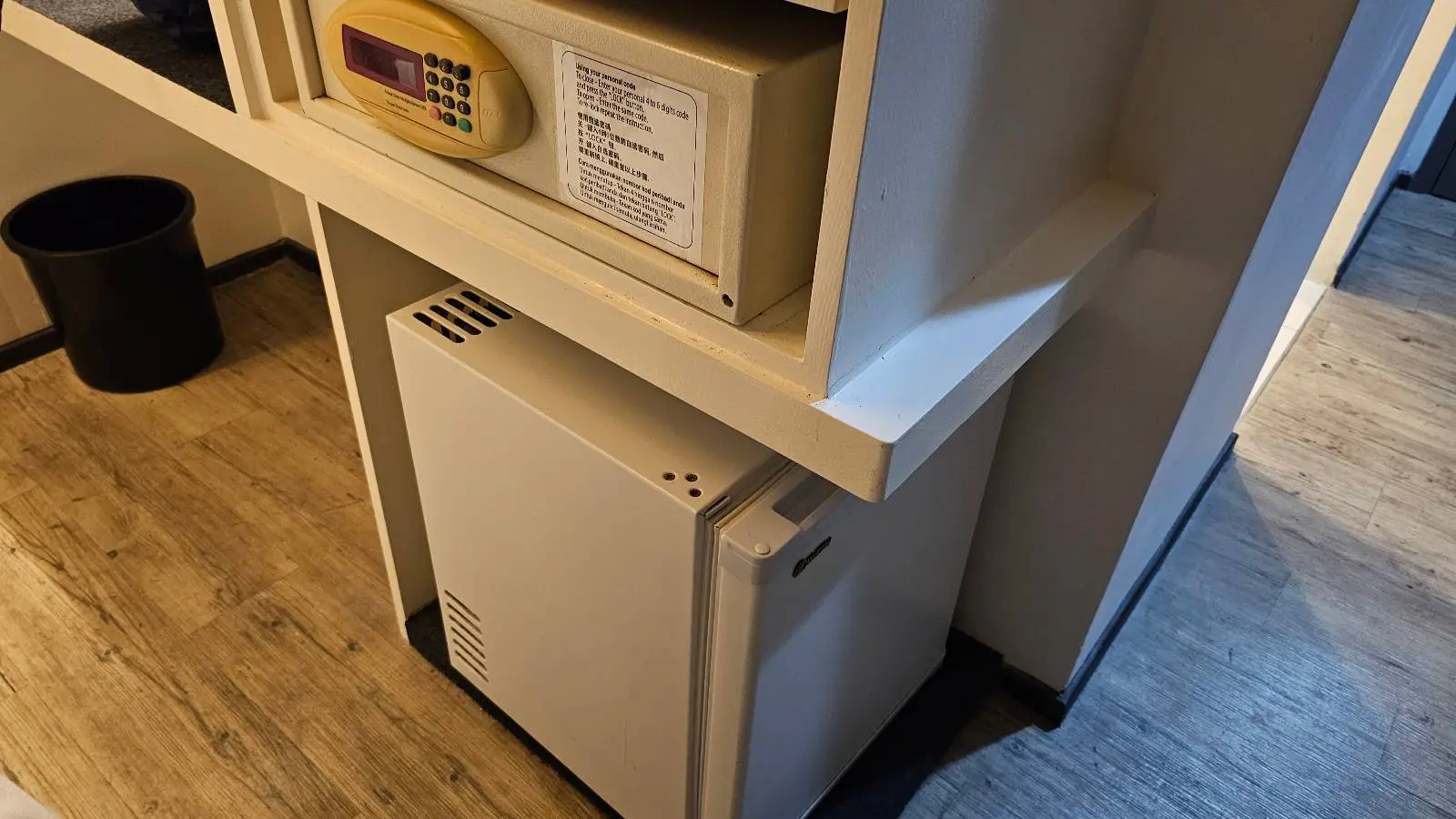 A small hotel room safe and a mini fridge are built into a white shelving unit against a wall, with a black trash bin on the wooden floor nearby.