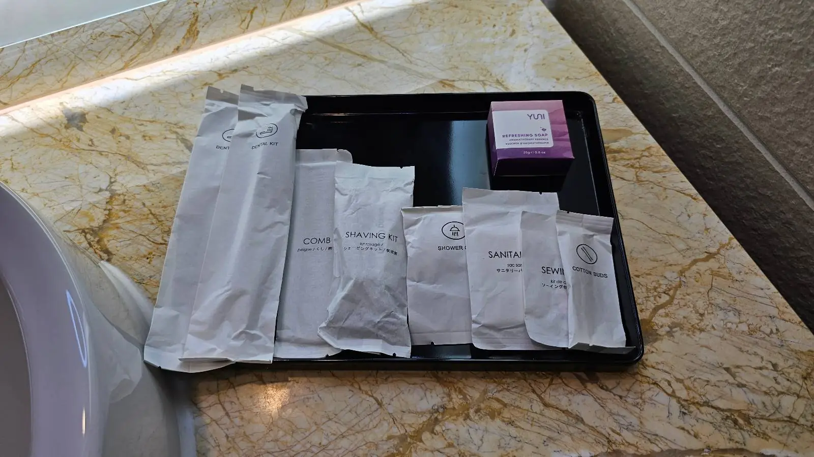 A tray on a marble countertop holds an assortment of neatly arranged white toiletries, including various packaged items and a small purple box, next to a bathroom sink.