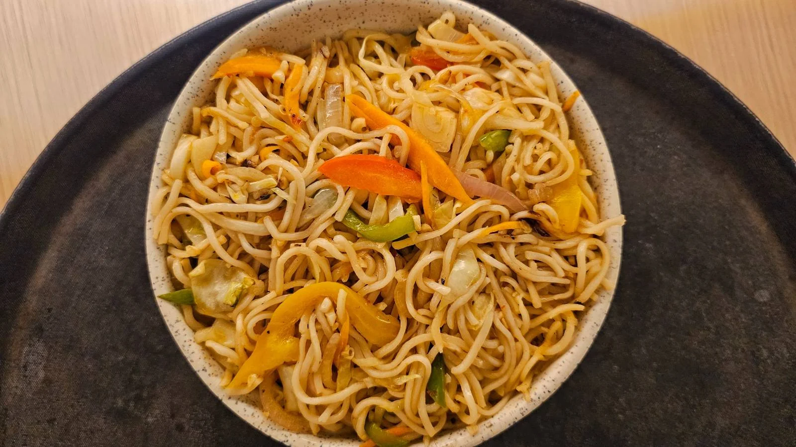 A bowl of stir-fried noodles with sliced carrots, bell peppers, and cabbage, served on a dark round tray on a wooden surface.