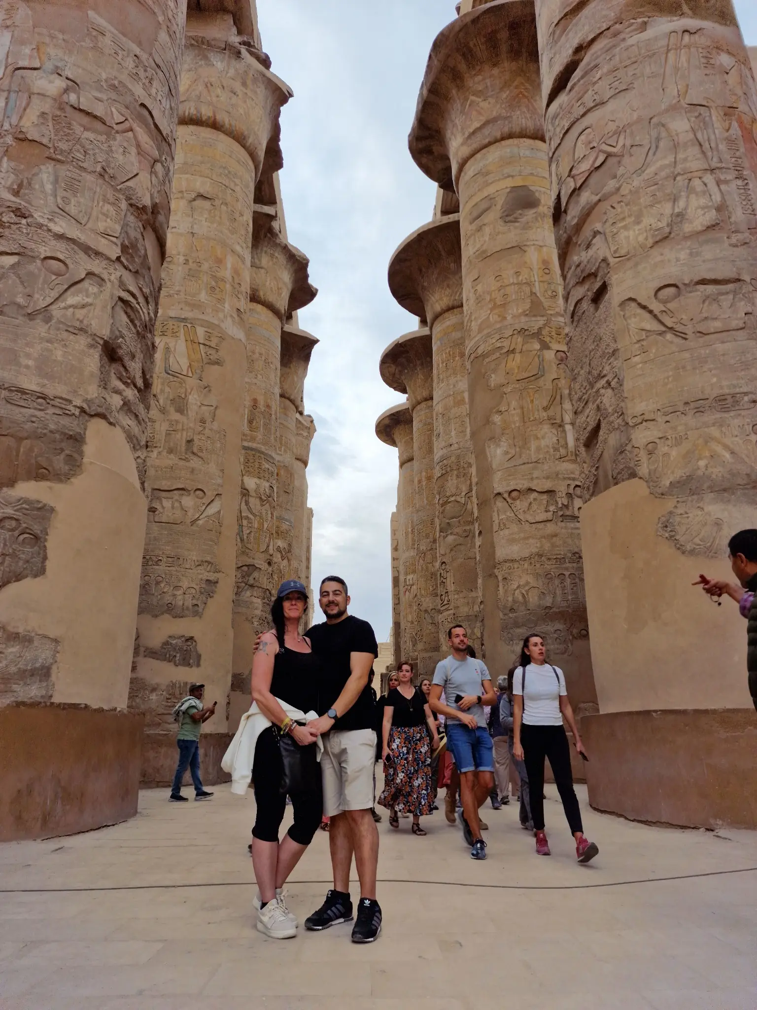 A group of people stands between towering ancient columns with intricate carvings. The sky is partly cloudy. The columns are weathered but grand, suggesting a historical or archaeological site.