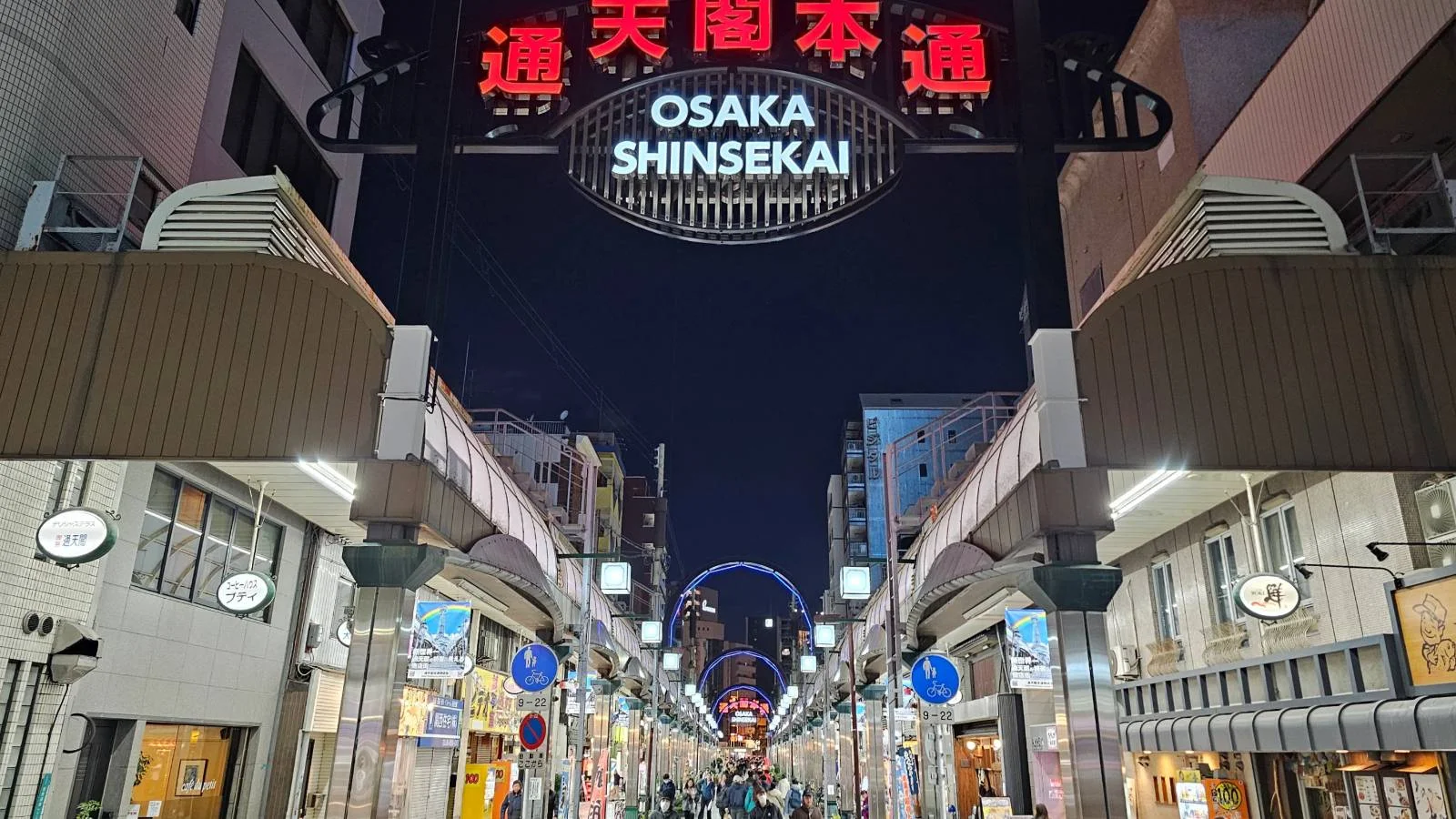 A brightly lit pedestrian street at night in Osaka, Japan. The entrance sign reads "Osaka Shinsekai" in English and Japanese, with shops and signs lining both sides of the street under a dark sky.