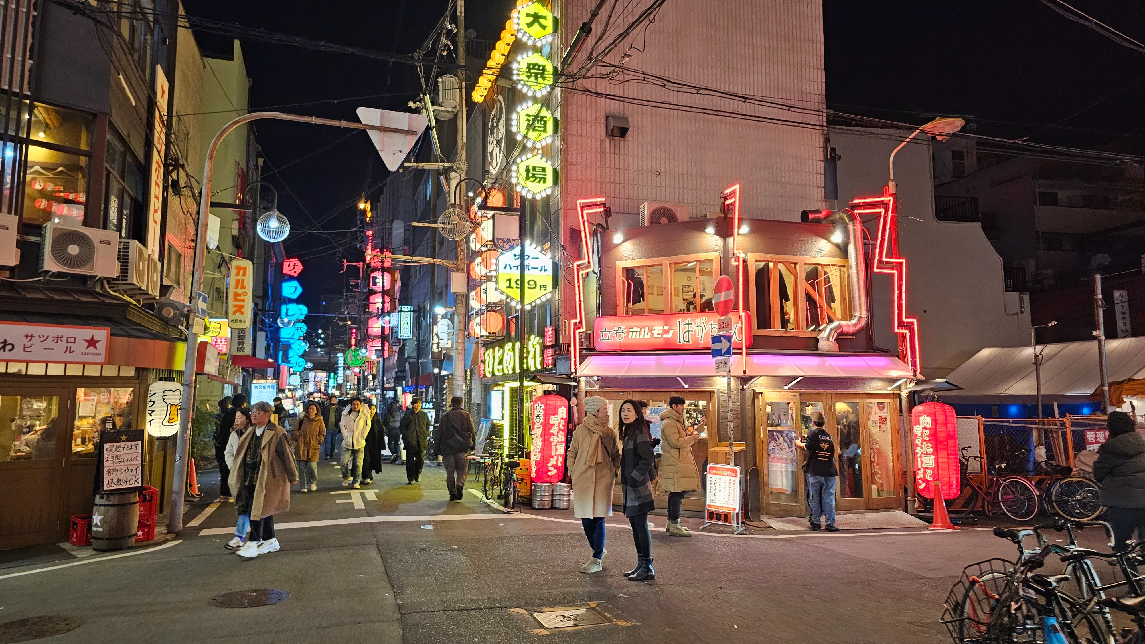 A lively city street at night with people walking, colorful neon signs, and lights illuminating shops and restaurants in an urban area.