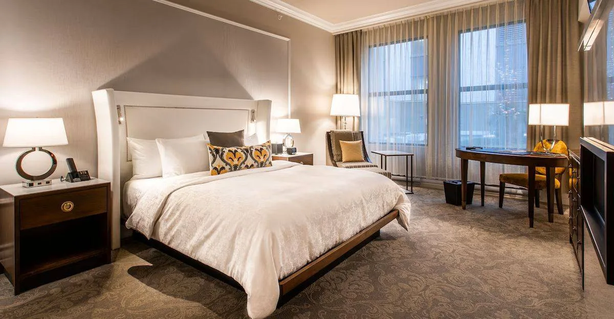 Elegant hotel room with a large bed, white linens, and patterned pillows. Warm lighting, carpeted floor, desk, and chair by sheer-curtained windows.
