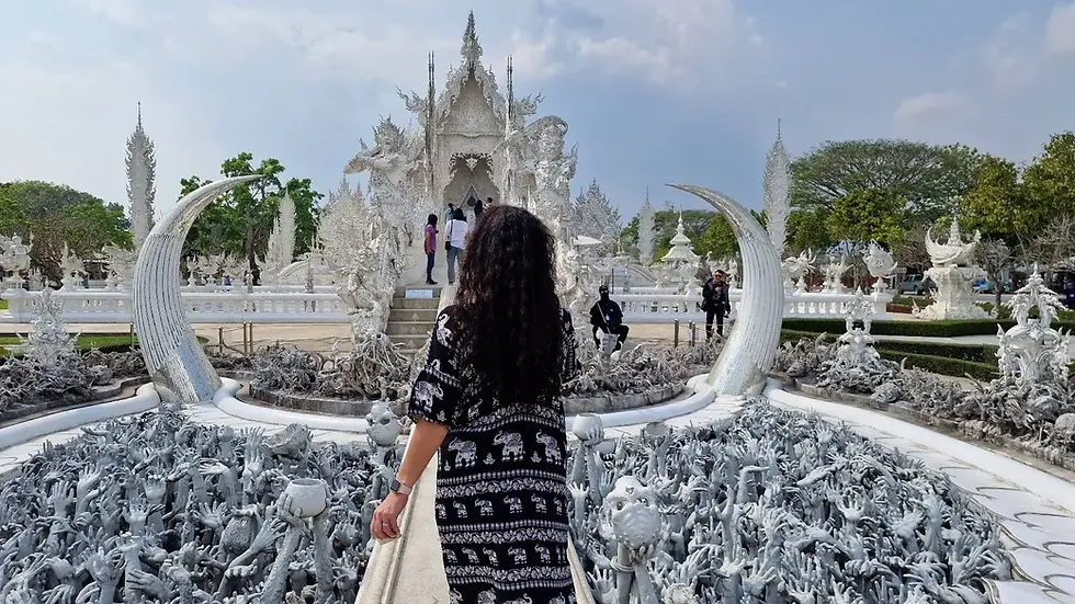 Woman in patterned dress walks across a bridge at a white ornate temple, surrounded by intricate sculptures and reaching hands, under a cloudy sky.