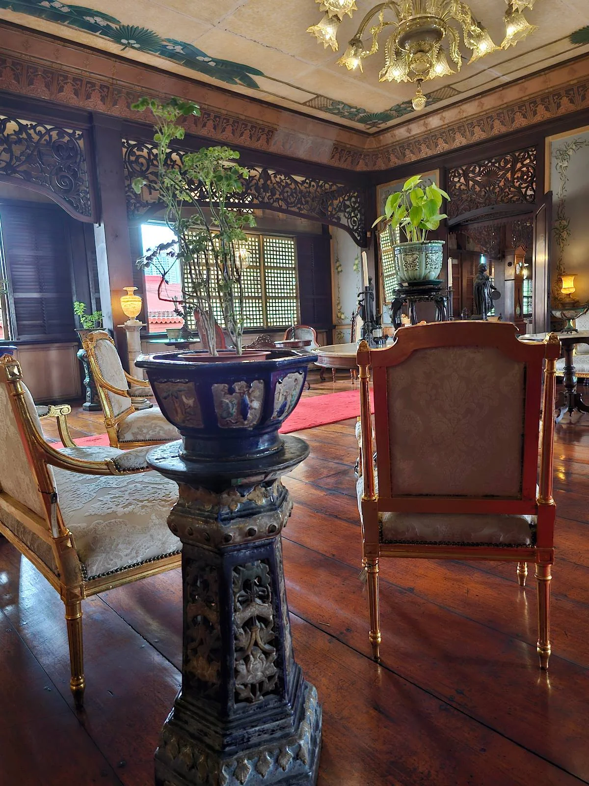 A luxurious room with ornate wooden walls and furniture, featuring elegant chairs and a large decorative plant on a pedestal. The high ceiling has intricate lighting, and a red rug adds warmth to the polished wooden floor.