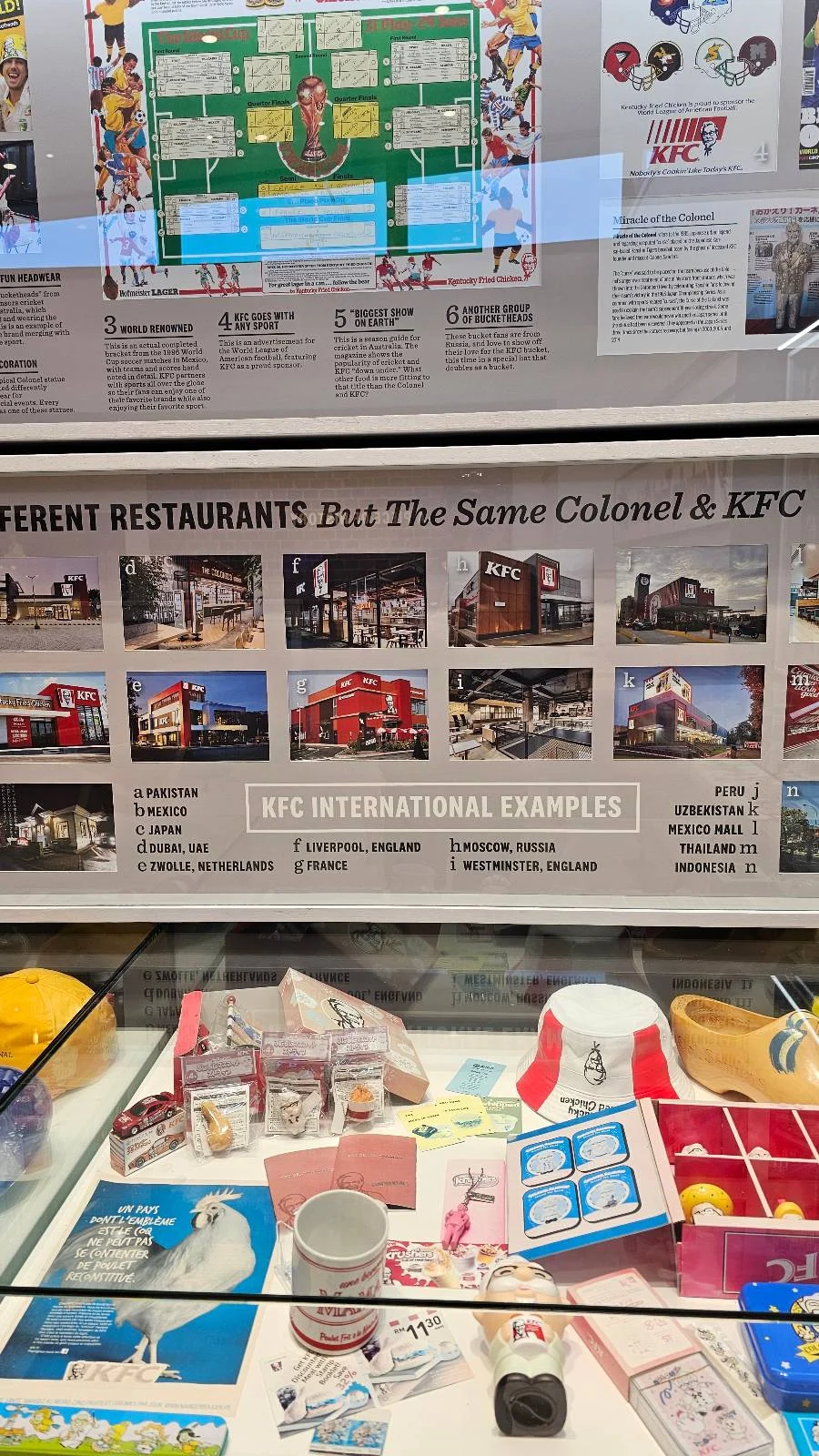 A museum display features memorabilia including old KFC packaging, restaurant photos, and a timeline about KFC’s history in the UK. Items include wrappers, cups, trays, and article clippings, all arranged behind glass.