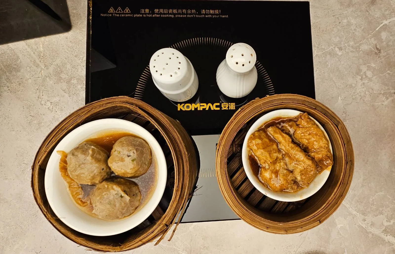 Two dishes on a cooktop: one with meatballs in sauce, the other with marinated tofu. Salt and pepper shakers are placed behind the dishes. The dishes are set in wooden trays on a black surface.